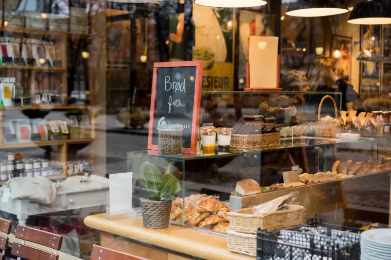 Google My Business optimieren - Eine Bäckereiauslage, die durch ein Fenster zu sehen ist. Auf einer Theke sind Brote, Gebäck und Gläser mit Waren angeordnet, perfekt für die Präsentation auf Google My Business. Auf einer Tafel steht „Brot“ mit einem unvollständigen Wort. Im Hintergrund sind Regale mit verschiedenen verpackten Artikeln zu sehen. Hängende Lichter beleuchten die Szene. Local SEO Agentur.