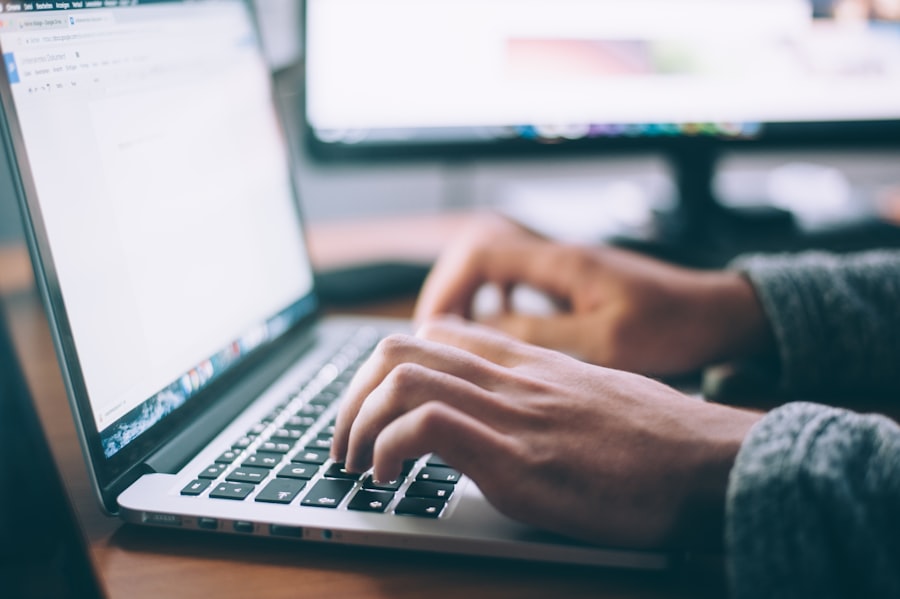 Google Unternehmensprofil Marketingstrategie 1 Google My Business optimieren - Eine Person tippt auf einer Laptop-Tastatur. Auf dem Bildschirm wird ein Dokument mit Text angezeigt. Ein weiterer Monitor im Hintergrund ist leicht unscharf. Die Umgebung scheint ein Schreibtischarbeitsplatz zu sein. Local SEO Agentur.