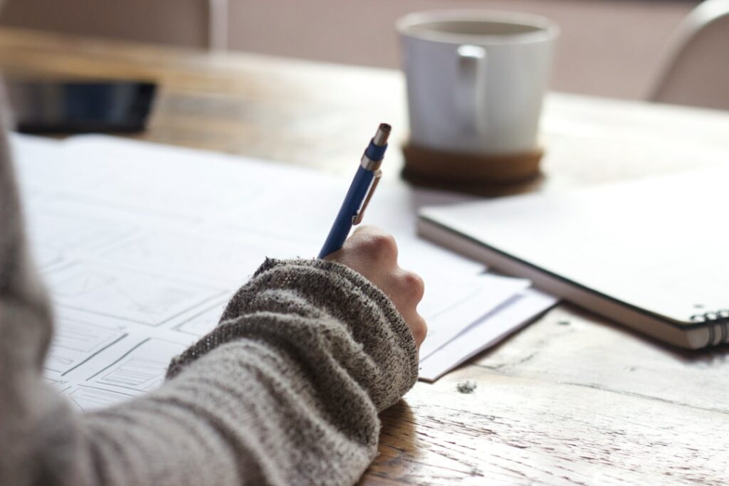 Google Unternehmensprofil optimieren lassen 7 google my business optimieren Die Hand einer Person hält einen Stift und schreibt auf einem Blatt Papier, das auf einem Holztisch liegt. Ein Notizblock und eine weiße Kaffeetasse auf einem Untersetzer liegen ebenfalls auf dem Tisch, neben Informationen zur Google my Business Agentur. Die Person trägt ein graues Langarmshirt.