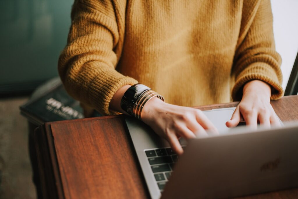Google My Business Eintrag verbessern 4 google my business optimieren Eine Person in einem senfgelben Pullover tippt auf der Tastatur eines Laptops, während sie an einem Holzschreibtisch sitzt. Das Gesicht der Person ist nicht sichtbar. Auf der Seite des Schreibtischs sind einige Bücher teilweise sichtbar.