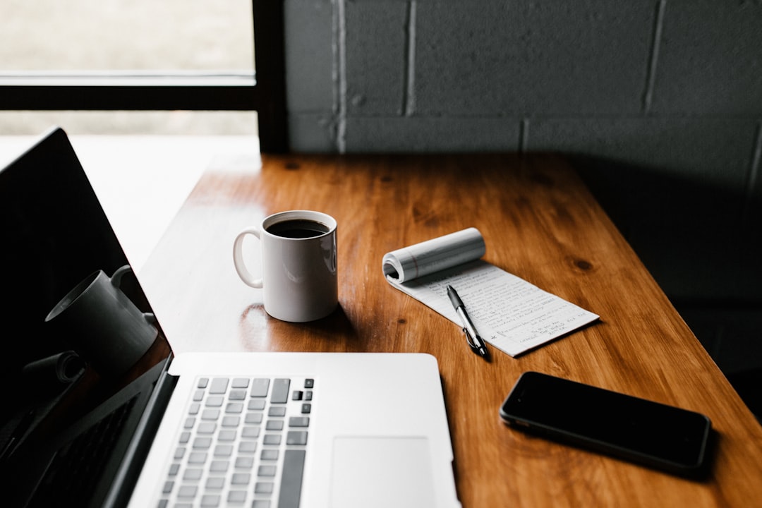 Google My Business optimieren - Auf einem Holzschreibtisch stehen ein Laptop, eine weiße Tasse mit Kaffee, ein Notizblock mit Schrift, ein Stift und ein Smartphone. Der Schreibtisch steht neben einem Fenster, so dass die Gegenstände von natürlichem Licht beleuchtet werden. Local SEO Agentur.