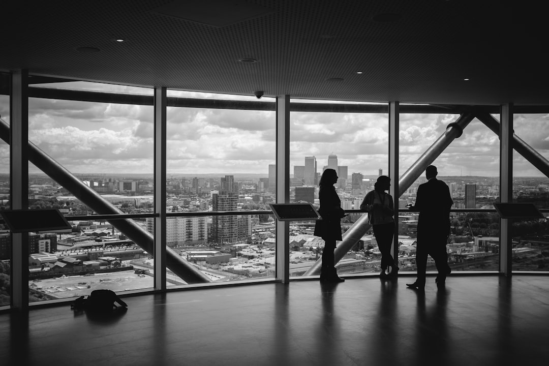 Google My Business optimieren - Drei Personen stehen und unterhalten sich vor großen Fenstern, die einen Panoramablick auf die Skyline einer Stadt mit verschiedenen Gebäuden bieten. Die Szene scheint sich in einem Innenbereich zu befinden, möglicherweise in einem Büro oder auf der Aussichtsplattform eines Hochhauses. Das Bild ist schwarzweiß. Local SEO Agentur.